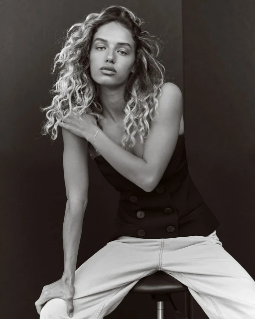 Portrait of a woman with curly blonde hair, wearing a black top and light jeans, sitting on a stool in front of a black backdrop.