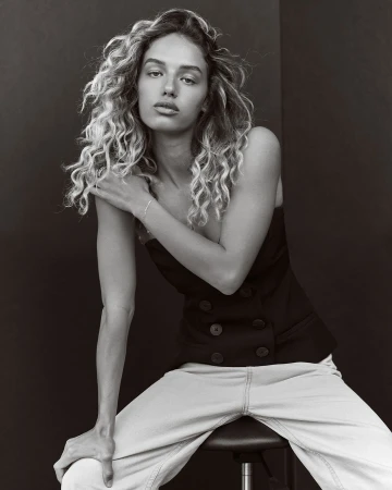 Portrait of a woman with curly blonde hair, wearing a black top and light jeans, sitting on a stool in front of a black backdrop.
