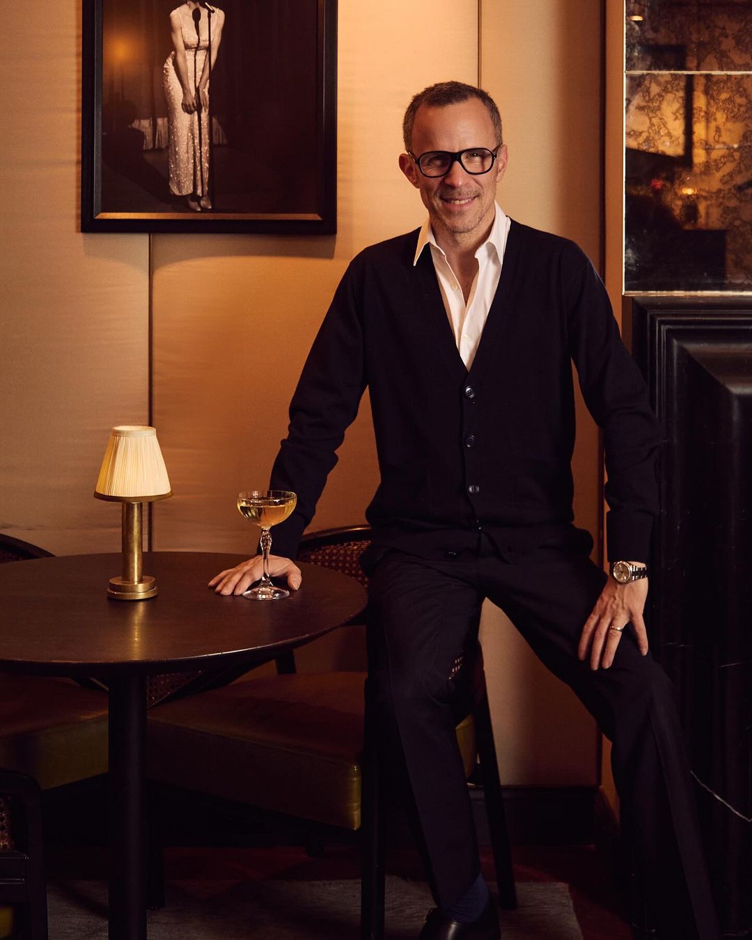 Elegant man in glasses sits near a table with a cocktail, wearing a black cardigan and pants, posing in a warm, intimate setting.