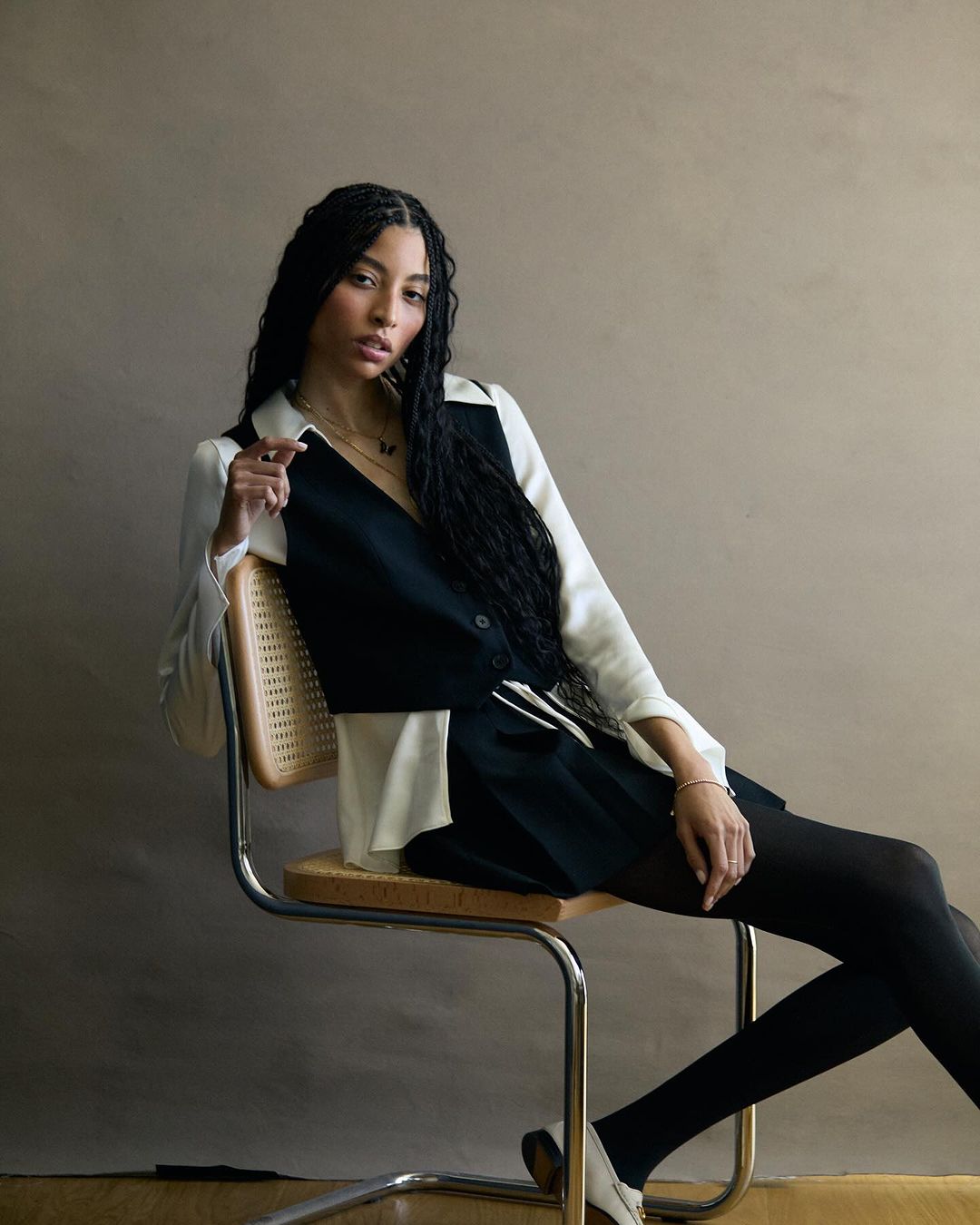 Stylish woman with braided hair sits on a chair in a two-tone black and white outfit, completed with stockings and heels against neutral backdrop.