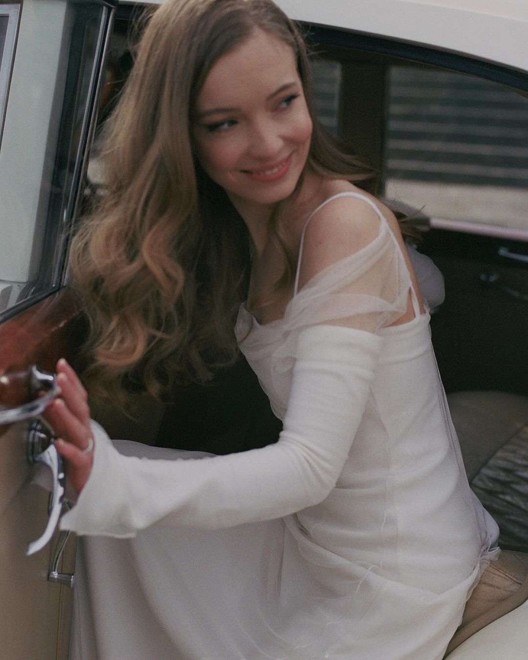 Blonde woman in a white dress exiting a vintage car with a smile, long sleeves, tulle details, holding the door.