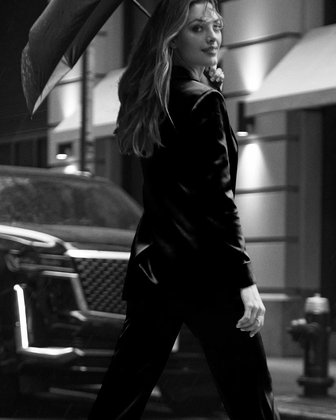 Black and white portrait of a woman in a suit, holding an umbrella, walking on a city street with car and buildings in the background.