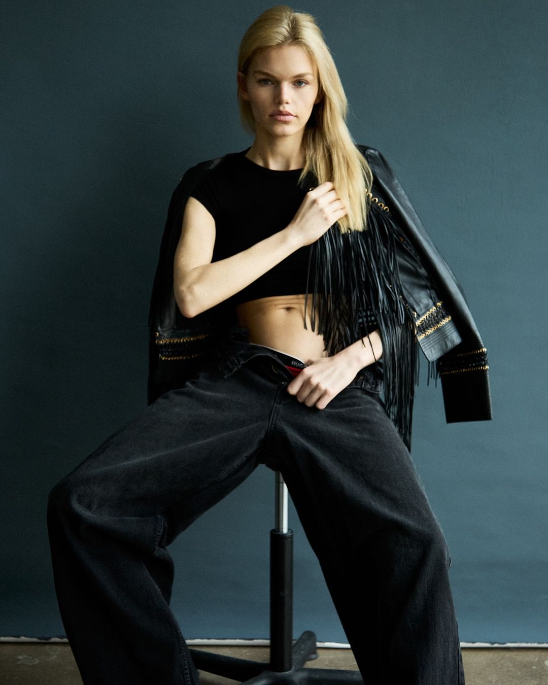 Blonde model sits on a stool wearing a black crop top, wide-leg jeans, and a fringed jacket against a dark blue background in a studio shot.