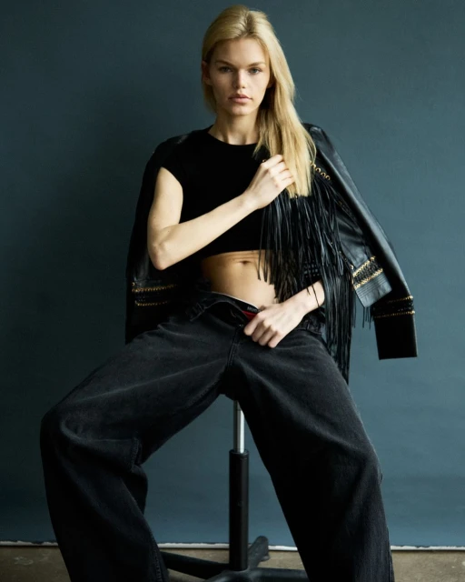 Blonde model sits on a stool wearing a black crop top, wide-leg jeans, and a fringed jacket against a dark blue background in a studio shot.
