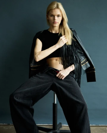 Blonde model sits on a stool wearing a black crop top, wide-leg jeans, and a fringed jacket against a dark blue background in a studio shot.