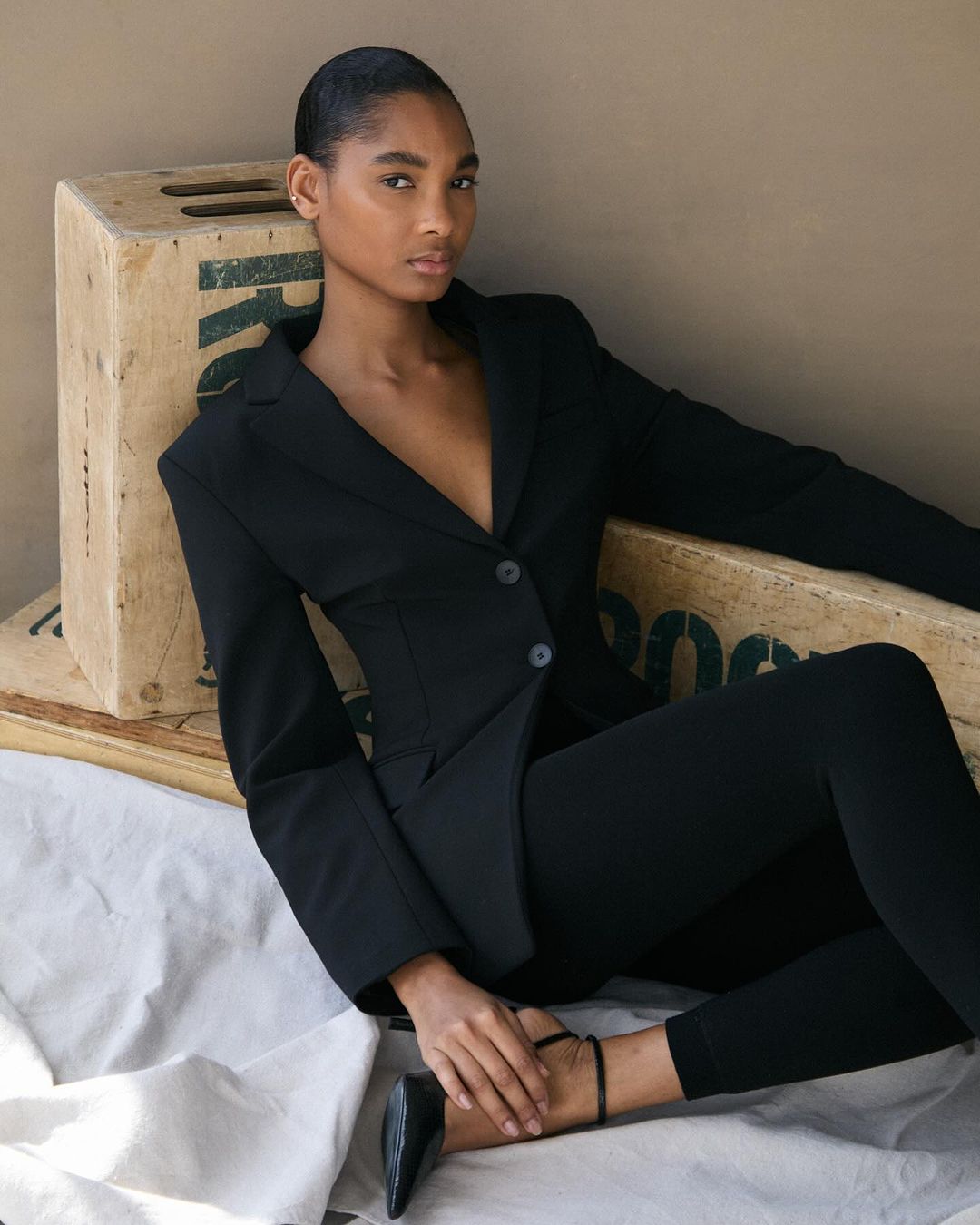 A young woman poses in a black blazer and pants while seated on wooden crates against a neutral backdrop in a fashion-style shot.
