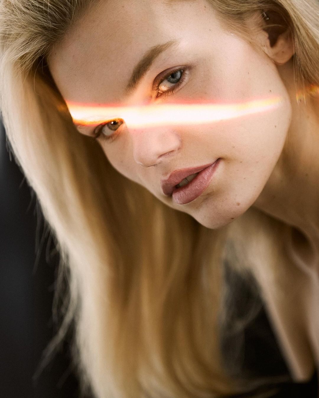 Close-up of a fair-skinned adult woman with light blonde hair, angled head, and a bar of light across her face against a dark background.