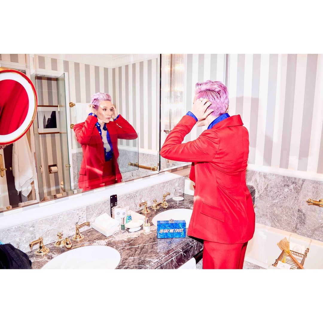 Woman in a red suit adjusts her hair in a marble-countered bathroom mirror with striped wallpaper, bright lighting, and vanity items.