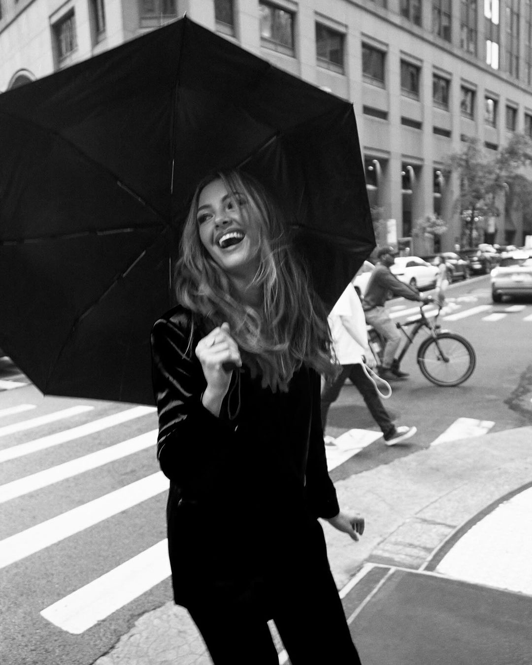 A woman laughs under an umbrella in a black and white photo, walking on a crosswalk in an urban environment with buildings behind her.