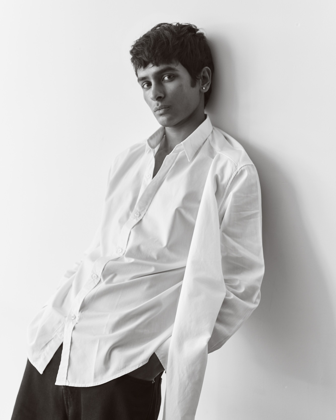 Studio portrait of a young adult leaning against a white wall, wearing a white dress shirt and dark pants in black and white.