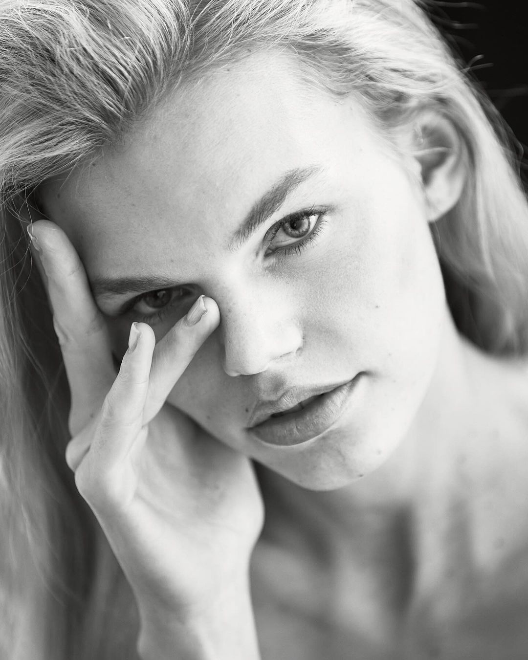 Monochrome portrait of a young adult woman with long blonde hair and fair skin, posing with her hand near her face.