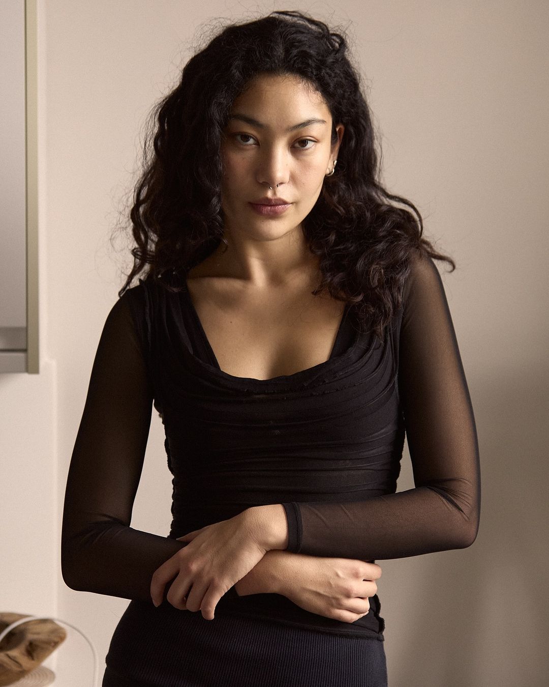 Portrait of an adult woman with dark curly hair, wearing a sheer black top, posing indoors in front of a neutral-toned wall.