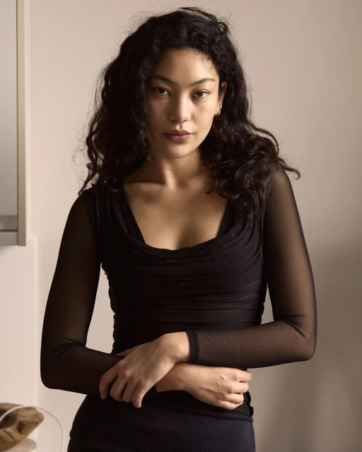 Portrait of an adult woman with dark curly hair, wearing a sheer black top, posing indoors in front of a neutral-toned wall.