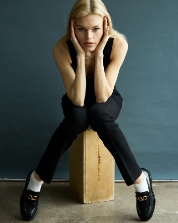 Blonde female model seated on a wooden crate wearing black trousers and loafers photographed by Andrew Day
