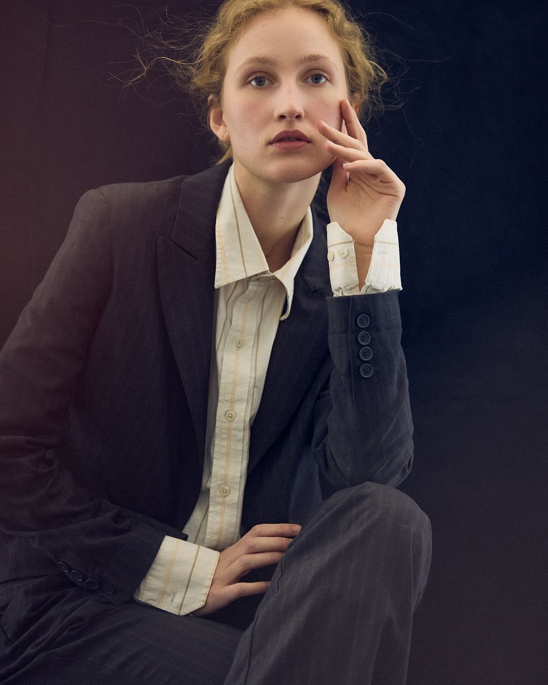 Young woman in a dark business suit and striped shirt poses in a studio portrait against a dark background, looking directly at the camera.
