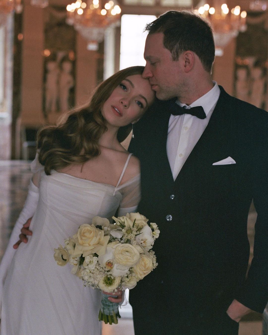 Romantic portrait of a bride in a white wedding dress with a bouquet, leaning against the groom in a black suit in an ornate hall.