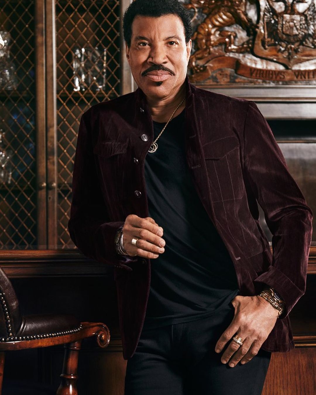 Close-up portrait of Lionel Richie wearing a burgundy jacket and black shirt, posing indoors against an ornate backdrop.