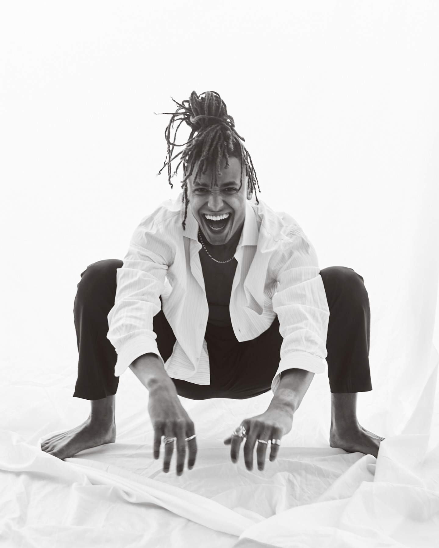 A joyful person with locs squats indoors on white fabric, smiling broadly, wearing a white shirt, black pants, and rings on their fingers.