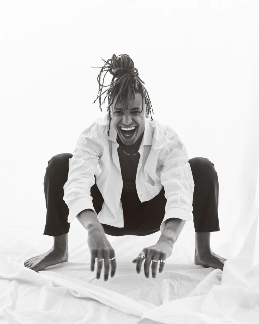 A joyful person with locs squats indoors on white fabric, smiling broadly, wearing a white shirt, black pants, and rings on their fingers.