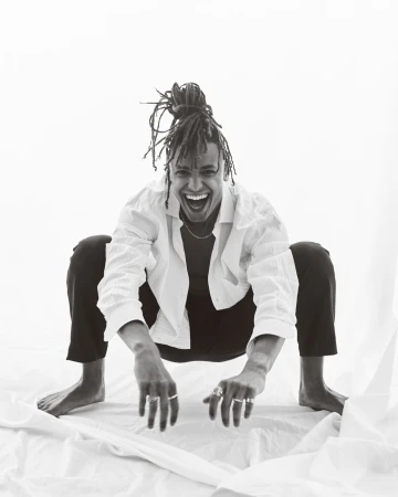 A joyful person with locs squats indoors on white fabric, smiling broadly, wearing a white shirt, black pants, and rings on their fingers.