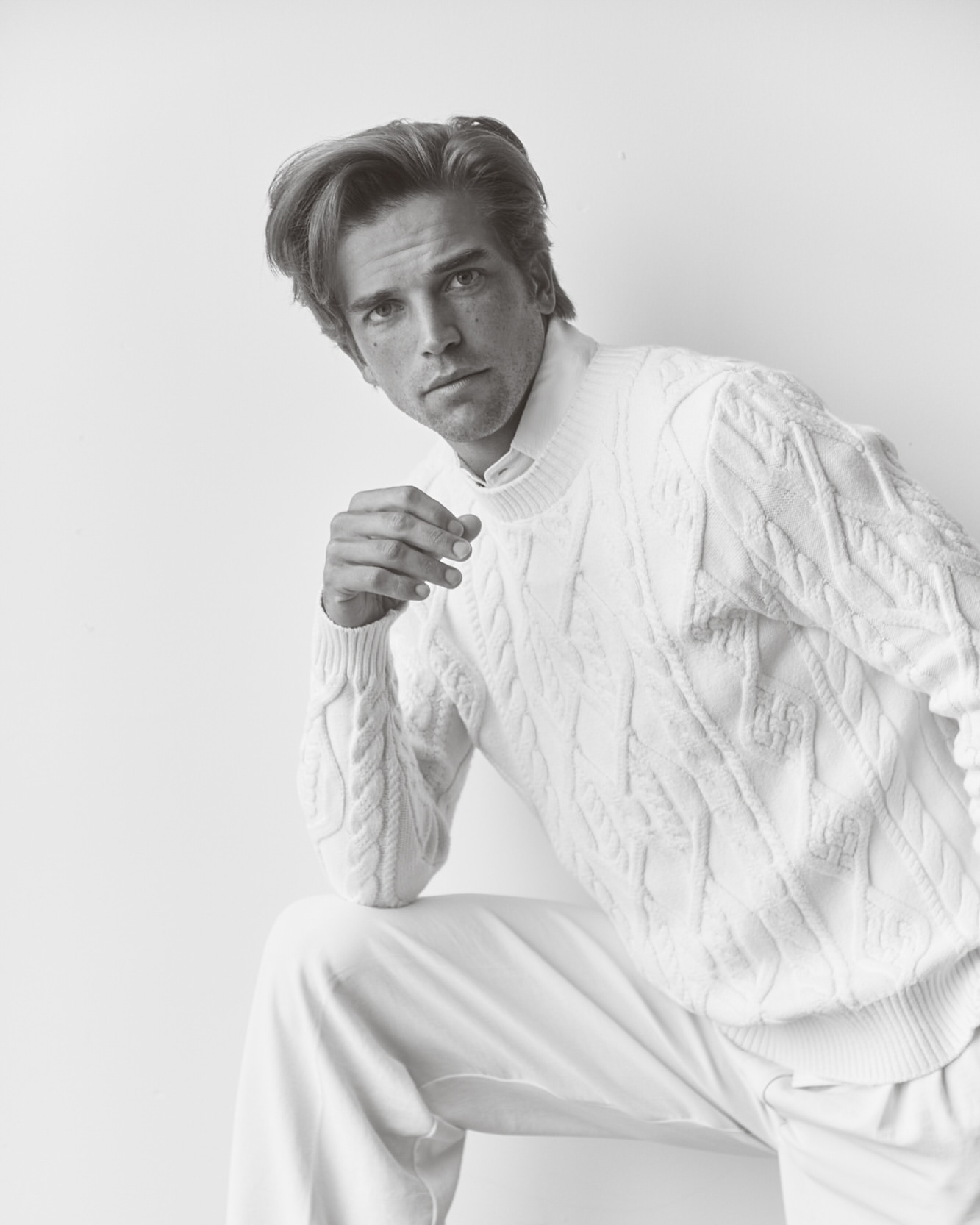 Monochrome portrait of a blond male model in a white cable-knit sweater and pants against a white backdrop in a studio setting.
