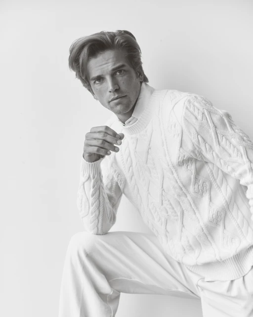 Monochrome portrait of a blond male model in a white cable-knit sweater and pants against a white backdrop in a studio setting.