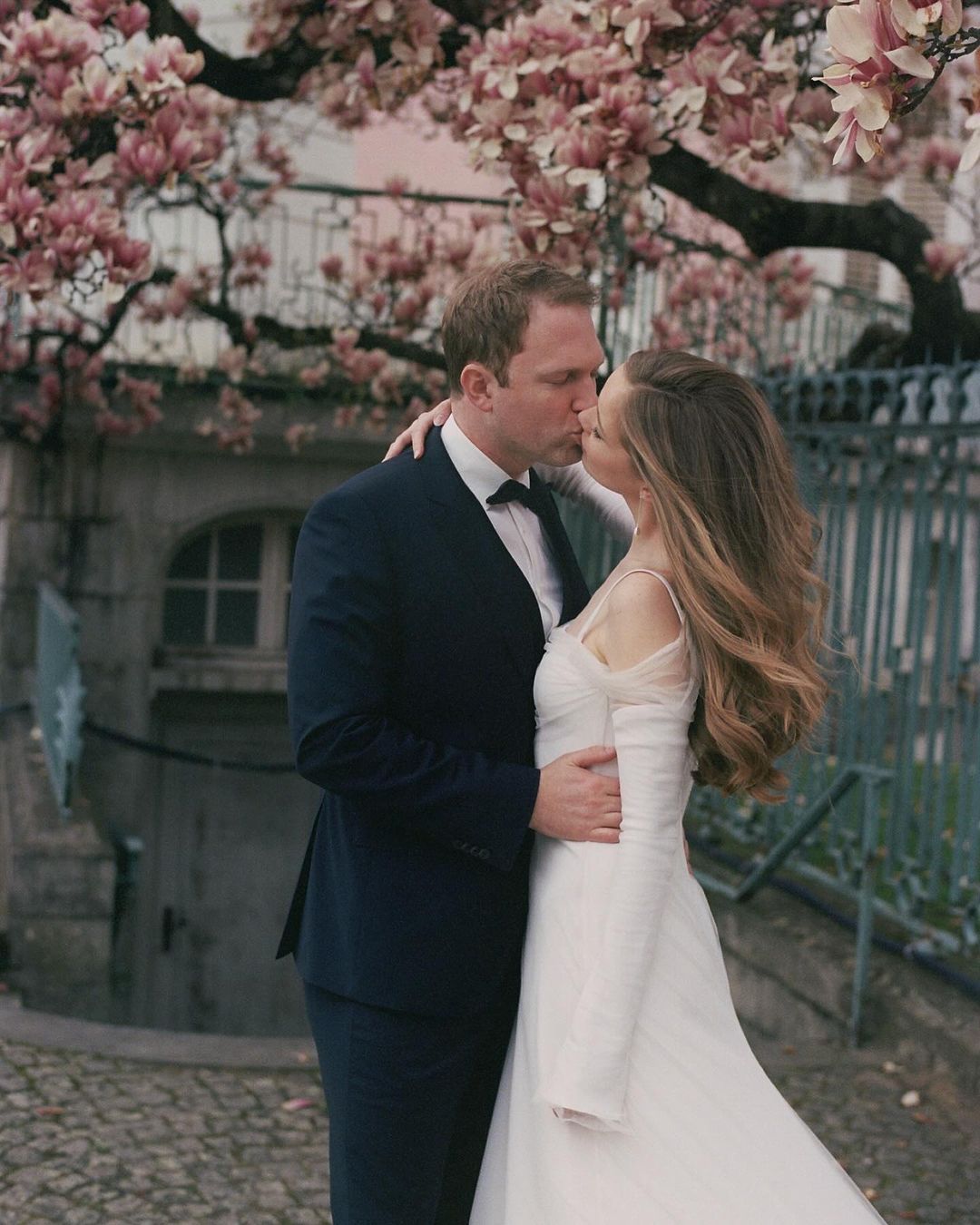 Elegant wedding photo of a couple kissing outdoors in front of a building with blossoming trees in soft, romantic light.