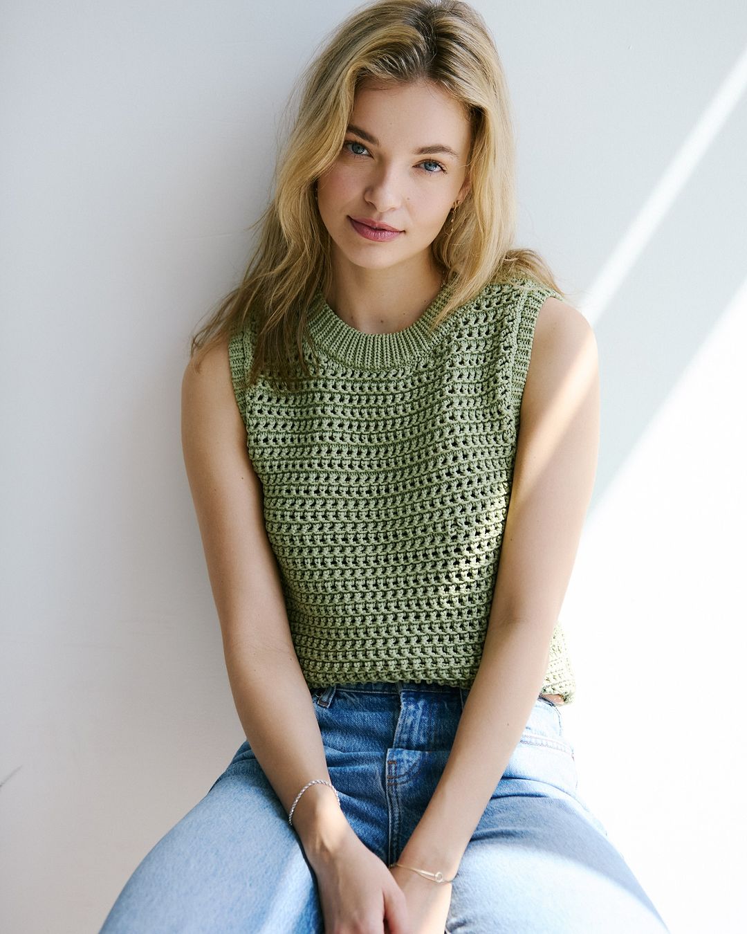 Blonde woman in an olive green crocheted tank top and blue jeans poses against a white wall with natural window light.