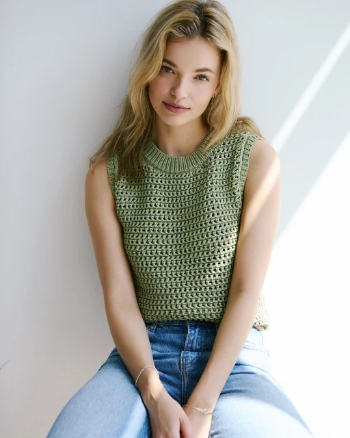 Blonde woman in an olive green crocheted tank top and blue jeans poses against a white wall with natural window light.