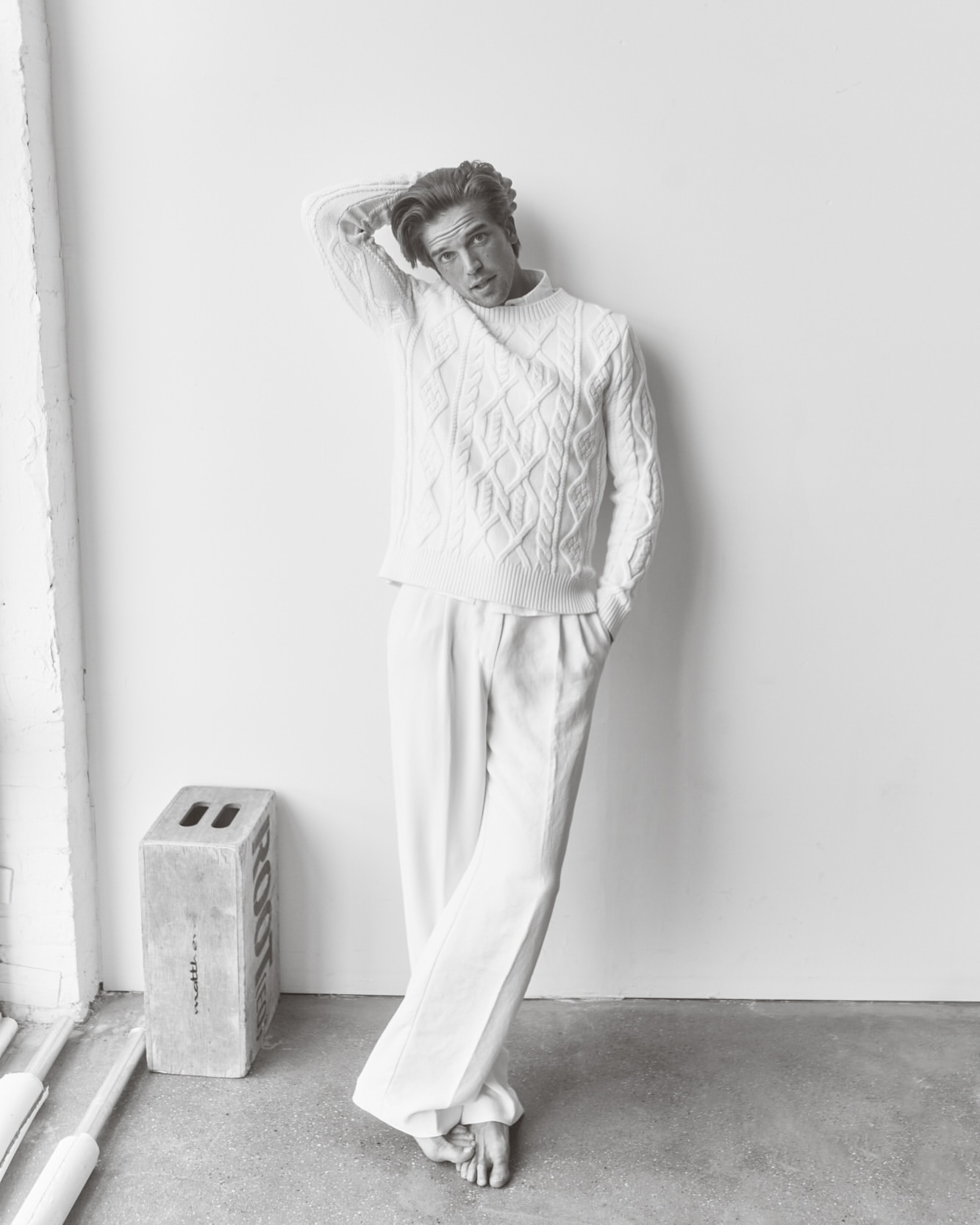 Monochromatic portrait of a young adult male model in a cable knit sweater and white pants, posing against a white wall in a photo studio.