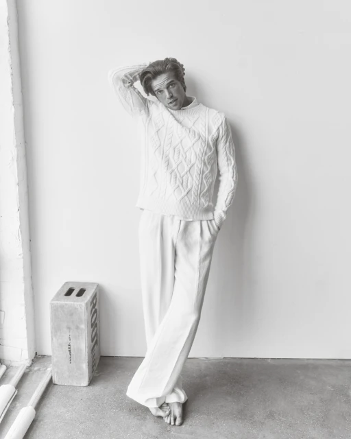 Monochromatic portrait of a young adult male model in a cable knit sweater and white pants, posing against a white wall in a photo studio.