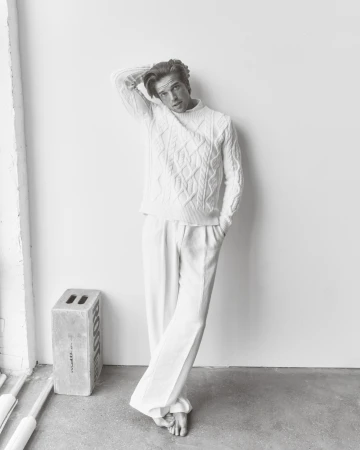 Monochromatic portrait of a young adult male model in a cable knit sweater and white pants, posing against a white wall in a photo studio.