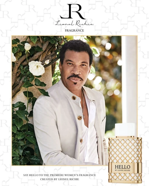 Lionel Richie poses in fragrance ad near white roses. The backdrop features a repeating R logo watermark pattern. He wears an open white jacket.