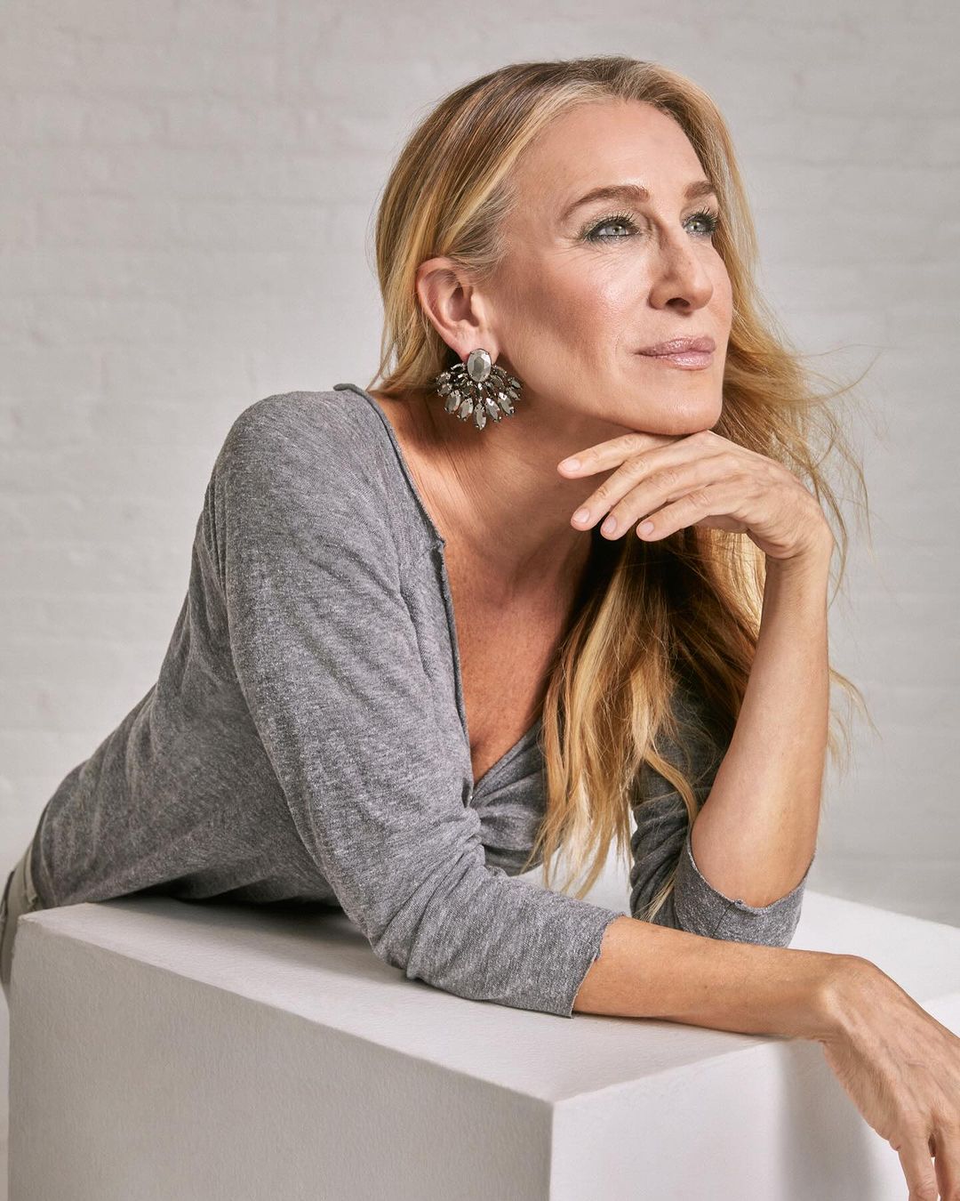 A portrait of a blonde woman with earrings, leaning on a white cube, in a photo studio with soft lighting and a white background.