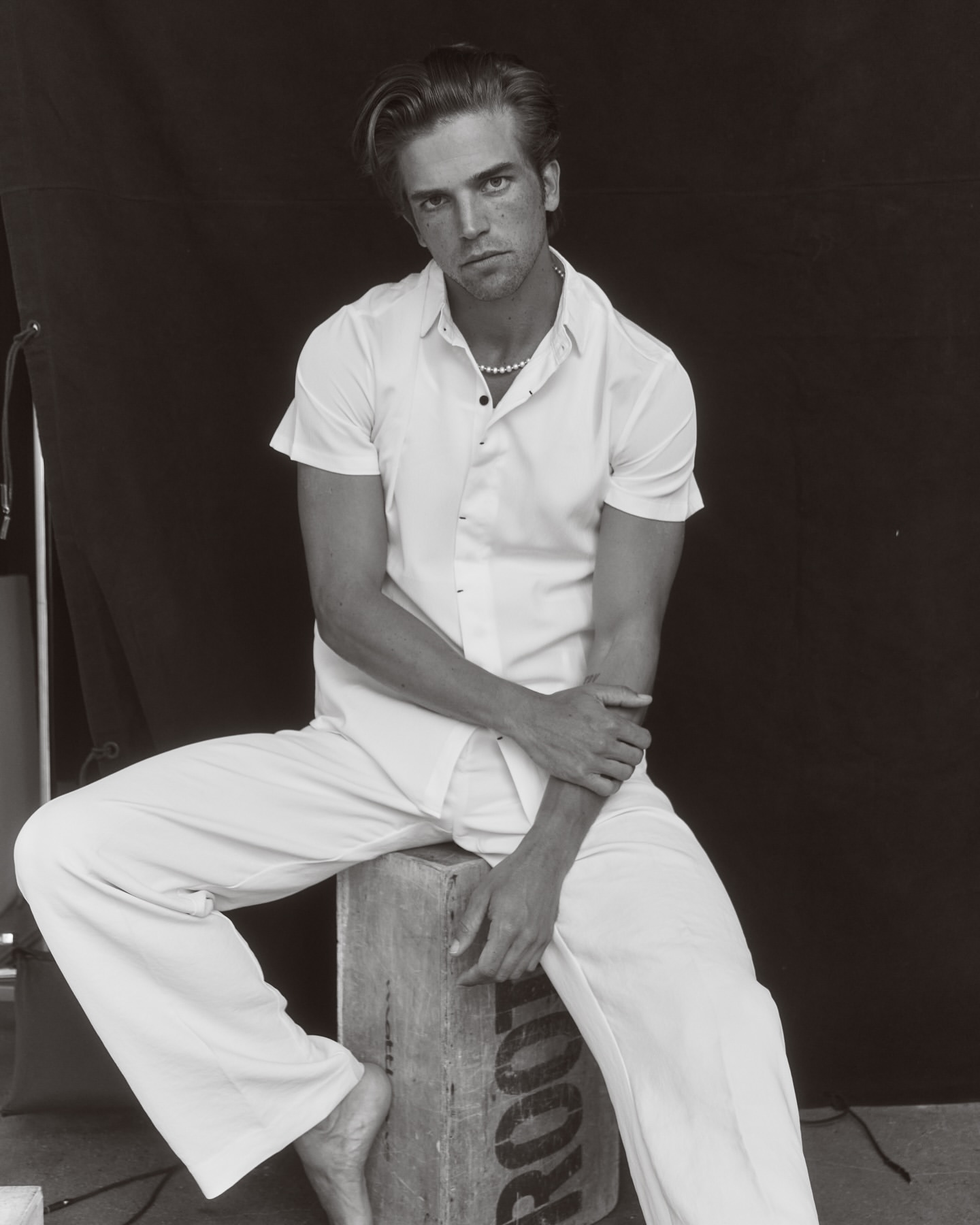 Monochrome portrait of a fair-skinned adult male in an unbuttoned white shirt and white pants seated on a wooden box in front of a black backdrop.