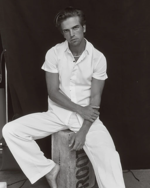 Monochrome portrait of a fair-skinned adult male in an unbuttoned white shirt and white pants seated on a wooden box in front of a black backdrop.