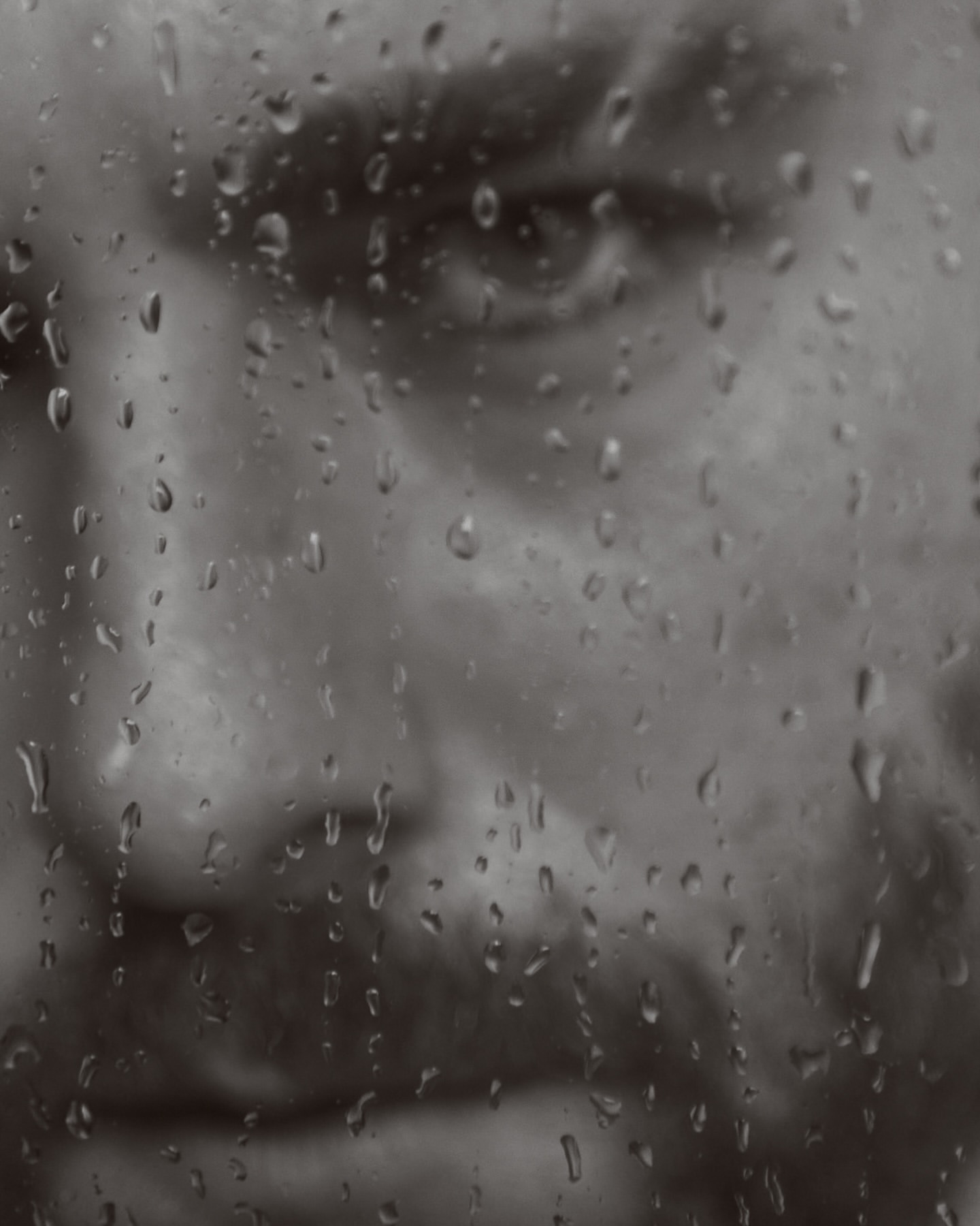 Monochrome portrait of an adult man's face behind a wet glass surface with raindrops, conveying a contemplative and melancholic mood.