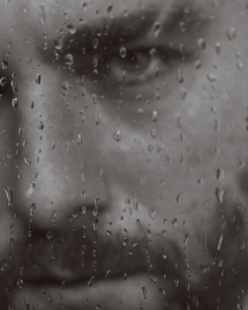 Monochrome portrait of an adult man's face behind a wet glass surface with raindrops, conveying a contemplative and melancholic mood.