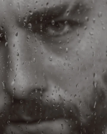 Monochrome portrait of an adult man's face behind a wet glass surface with raindrops, conveying a contemplative and melancholic mood.