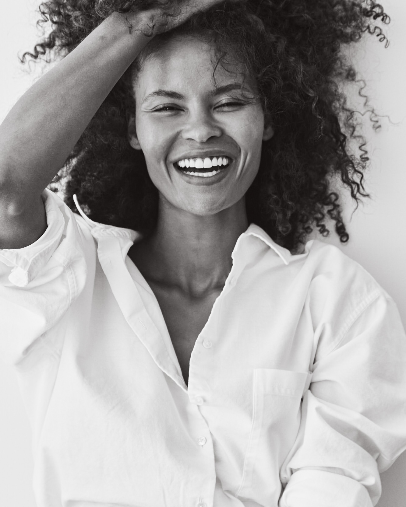Close-up studio portrait of a smiling adult female with curly hair, wearing a white collared shirt against a white backdrop.