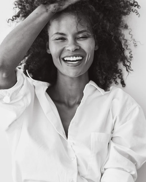 Close-up studio portrait of a smiling adult female with curly hair, wearing a white collared shirt against a white backdrop.