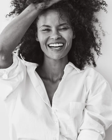 Close-up studio portrait of a smiling adult female with curly hair, wearing a white collared shirt against a white backdrop.