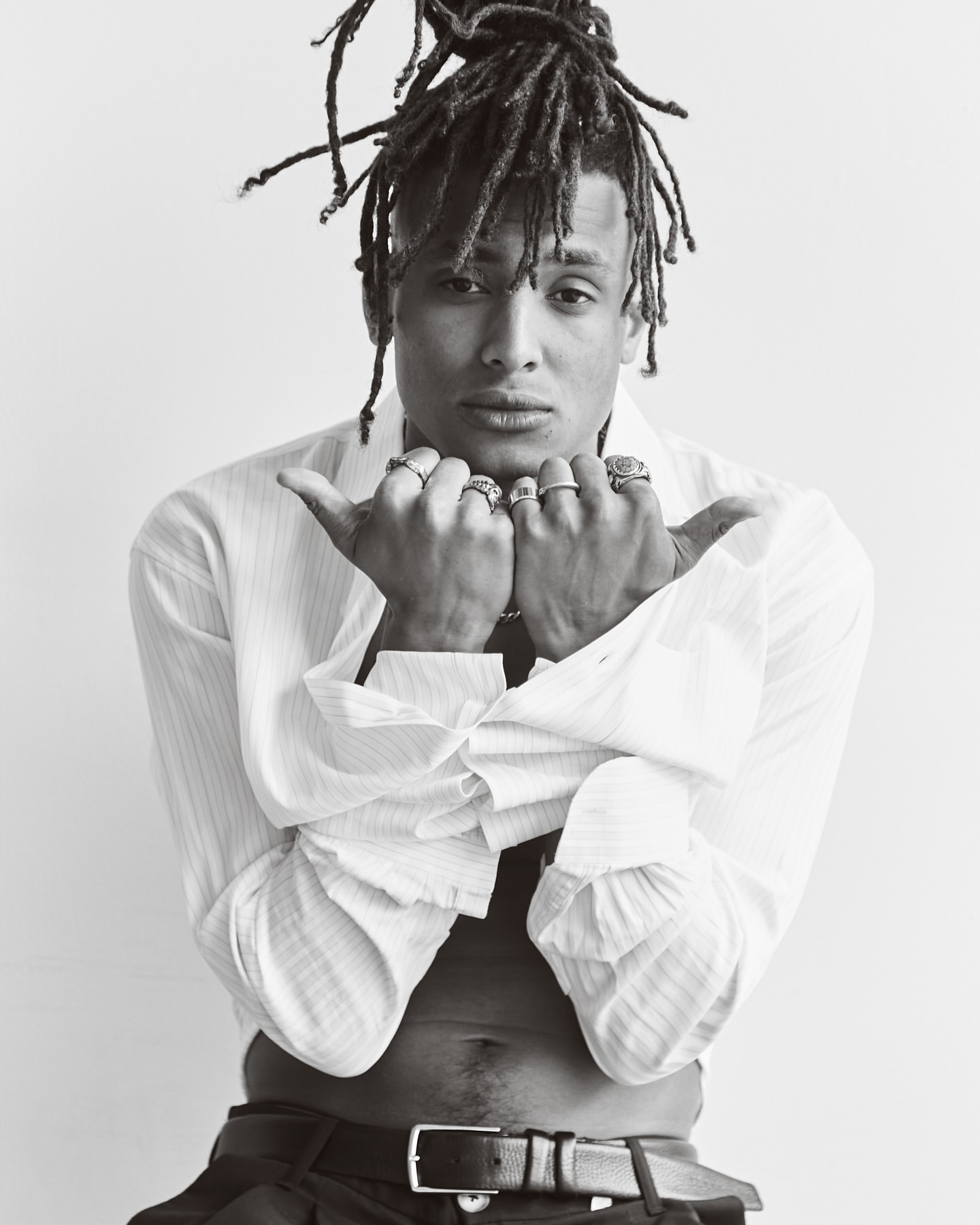 Monochrome portrait of a black man with dreadlocks wearing rings and an unbuttoned white shirt against a plain white background