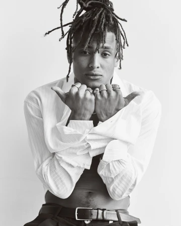 Monochrome portrait of a black man with dreadlocks wearing rings and an unbuttoned white shirt against a plain white background