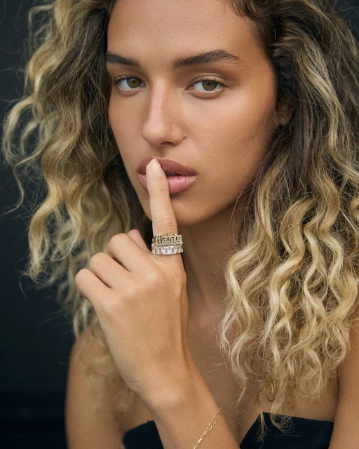 Portrait of a woman with blonde curly hair with a gold and diamond ring holding a finger up to her lips in front of a black background.