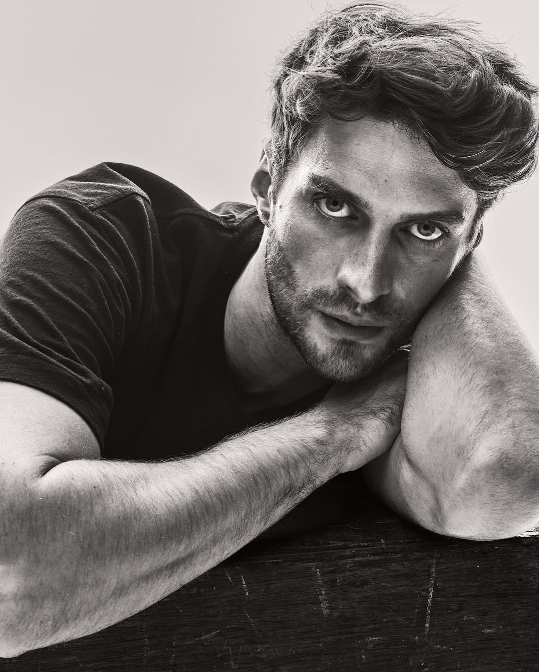 Black and white portrait of a handsome adult male resting his arms on a dark wooden surface, looking directly at the camera.