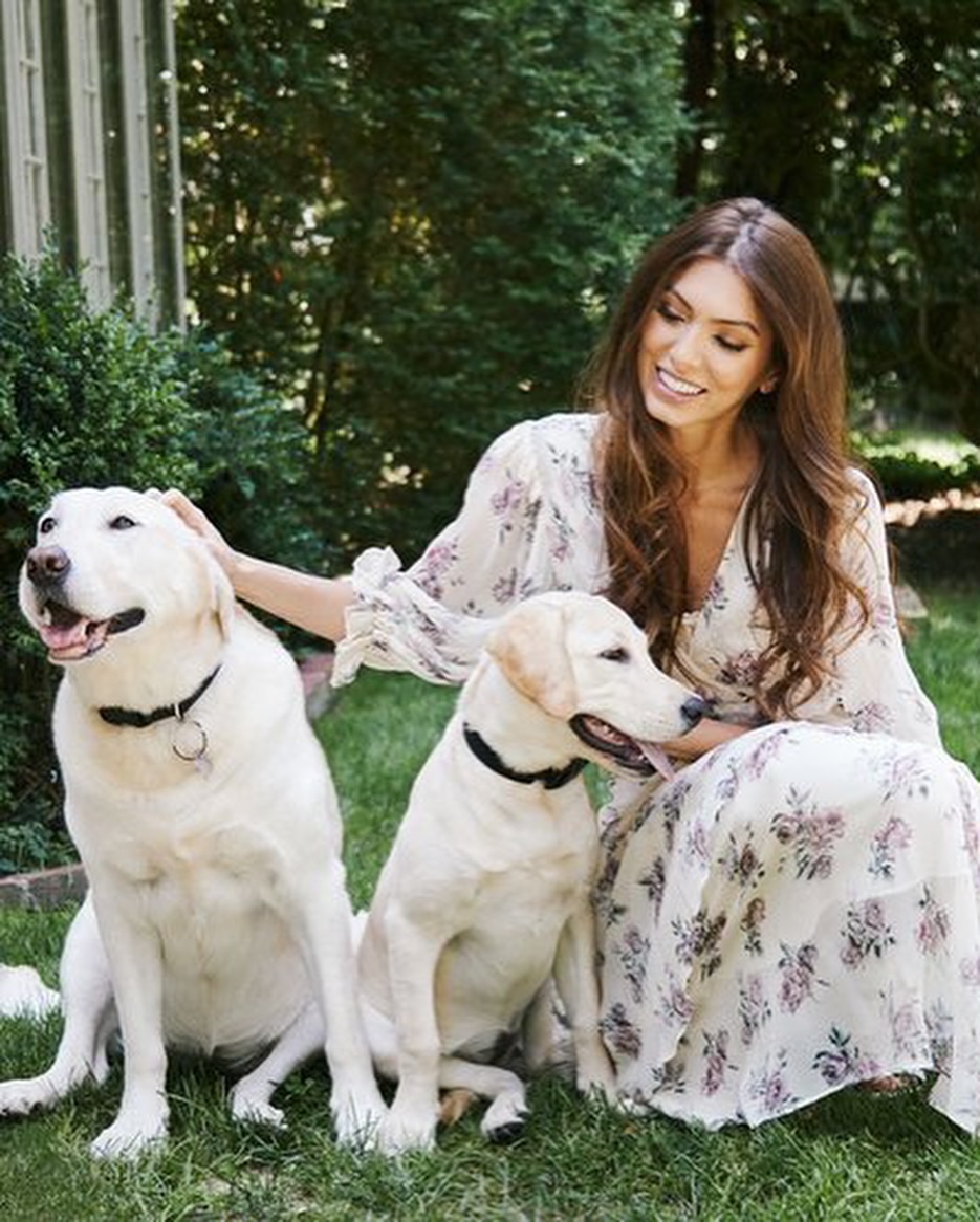 Woman in a floral dress poses outdoors with two Labrador Retriever dogs, sitting on the grass in front of lush green foliage.