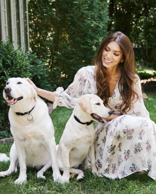 Woman in a floral dress poses outdoors with two Labrador Retriever dogs, sitting on the grass in front of lush green foliage.