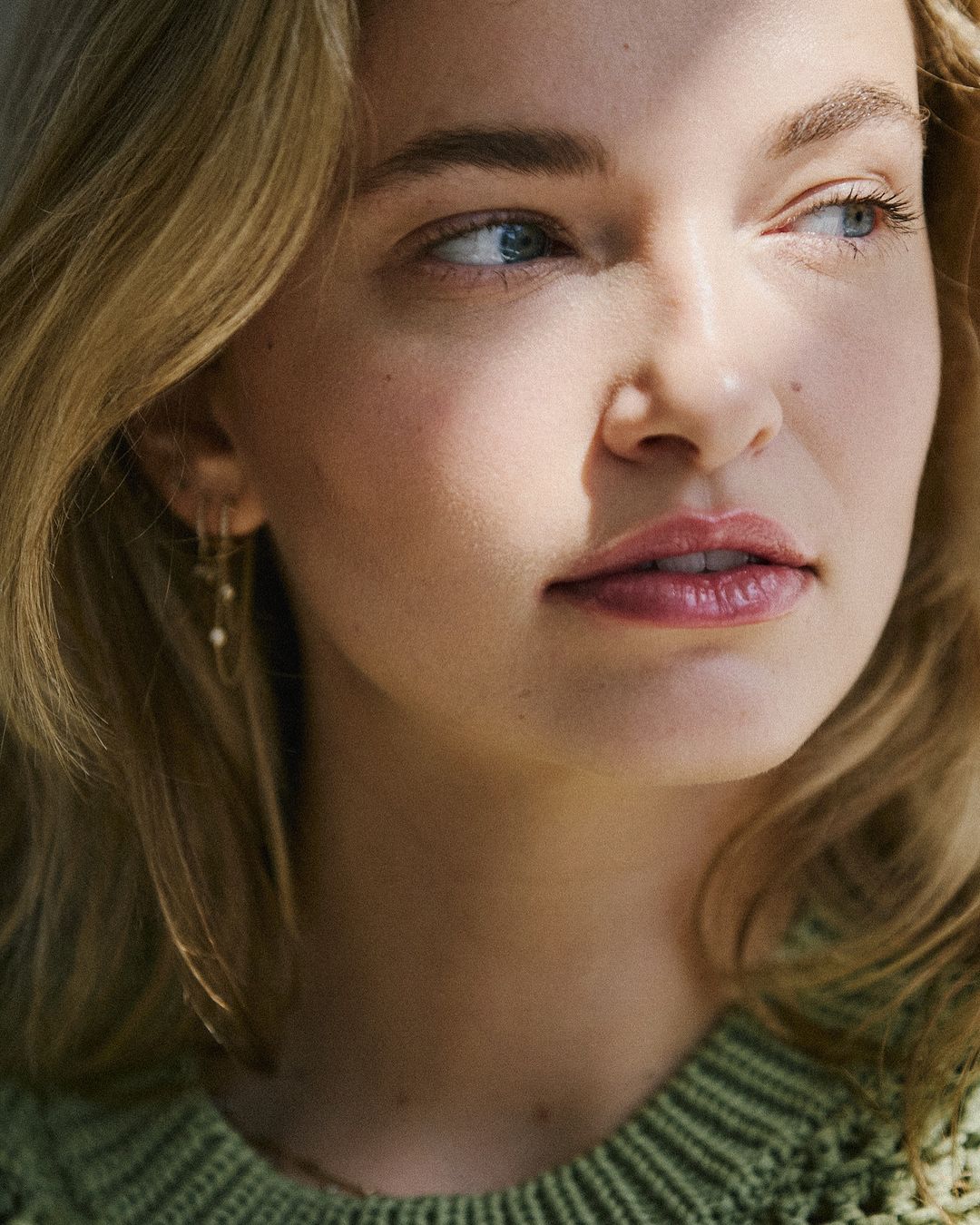 Close-up portrait of a woman with blonde hair wearing a green knitted sweater and delicate earrings, lit by natural sunlight.