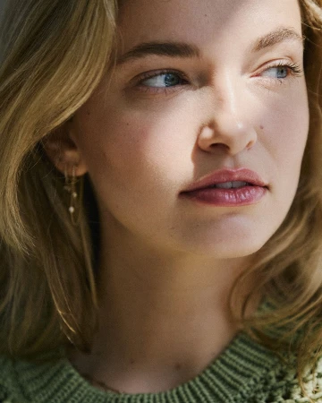 Close-up portrait of a woman with blonde hair wearing a green knitted sweater and delicate earrings, lit by natural sunlight.